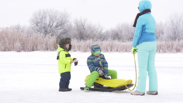 Mom in blue ski suit and three happy children in warm winter clothes ride on yellow sled on frozen snow-covered river.  Concept of friendly sports family. Winter holidays in village. Christmas eve