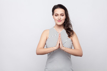 Portrait of beautiful young calm brunette woman with makeup and striped dress standing with closed eyes and palm hand in yoga pose and meditating. indoor studio shot, isolated on grey background.