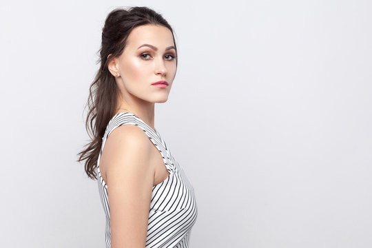 Profile Side View Portrait Of Serious Beautiful Young Brunette Woman With Makeup And Striped Dress Standing And Looking At Camera With Serious Face. Indoor Studio Shot, Isolated On Grey Background.