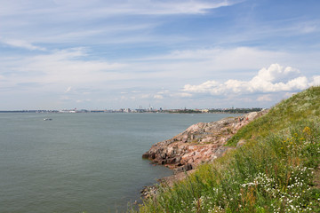 The stony shore of the island of Sumenlinna Sveaborg with grass and flowers on it and the view of the Gulf of Finland and the city of Helsinki in the distance on a summer day.