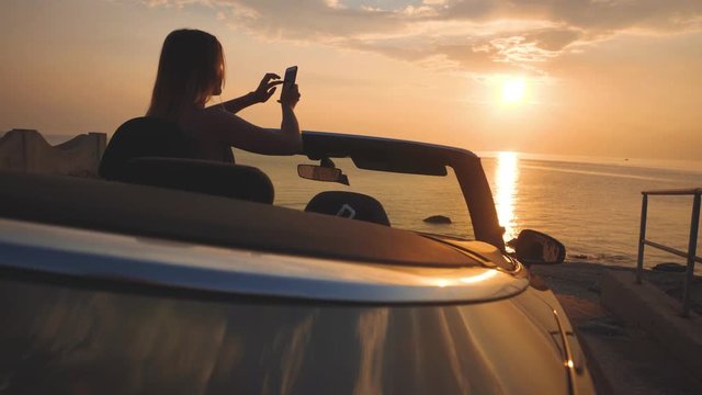 Girl takes a photo of sunset seascape by mobile phone from convertible car