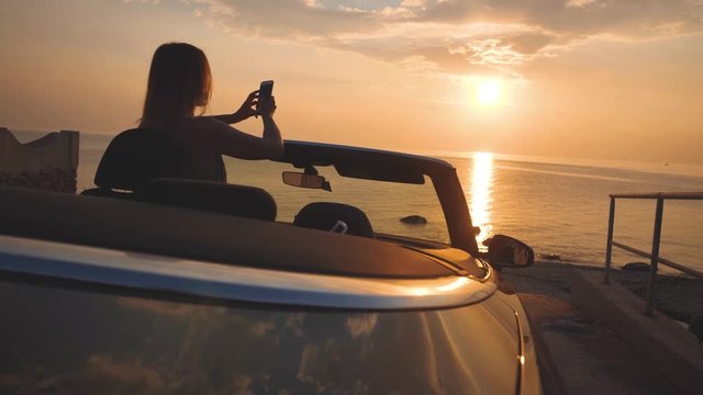 Female driver of luxury car takes s a photo of sunset seascape by cellphone