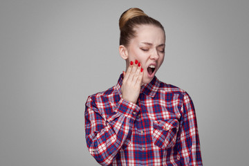 Tooth pain. unhappy blonde girl in red, pink checkered shirt, collected bun hairstyle, makeup standing touching her painful tooth with open mouth. indoor studio shot. isolated on gray background