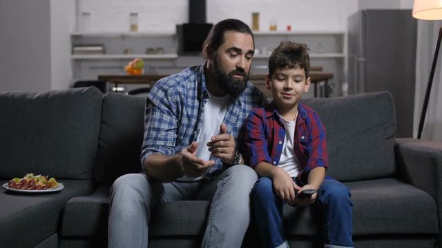 Handsome bearded stay-at-home father and preadolescent son watching tv and switching tv channels with remote control, unable to decide what to watch together while relaxing on sofa in domestic room