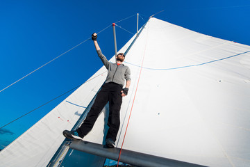 Nice and confident young man stands on yacht board and look at the sea through glasses. He is at the edge of yacht. Guy holding sail rope in his hand. Young man is happy and peaceful.