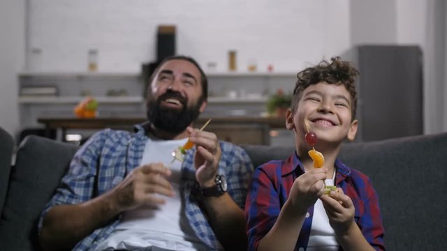 Portrait of carefree positive bearded father and smiling mixed race son eating fruit kebabs, chatting, enjoying freetime watching tv comedy show together in domestic room in the evening.