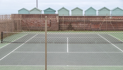 Tennis court, with row of green beach huts behind, on the sea front in Hove, Sussex, UK