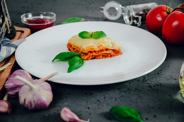 Lasagne on a white plate. On the stone table lies lasagna, spices and food ingredients. The concept of eating and preparing lasagna in tomato sauce.