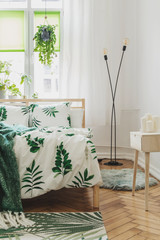Modern scandinavian sunny bedroom with plants , floral pattern bedding ,design lamp and pilows. Space with white walls and brown wooden parquet. 