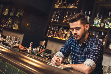 Young bartender leaning on bar counter taking notes concentrated