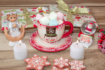 A mug with a snowman, marshmallow and gingerbread in the form of stars on a wooden table is decorated with fir branches, angels, candles and gifts. Happy New Year and Merry Christmas.
