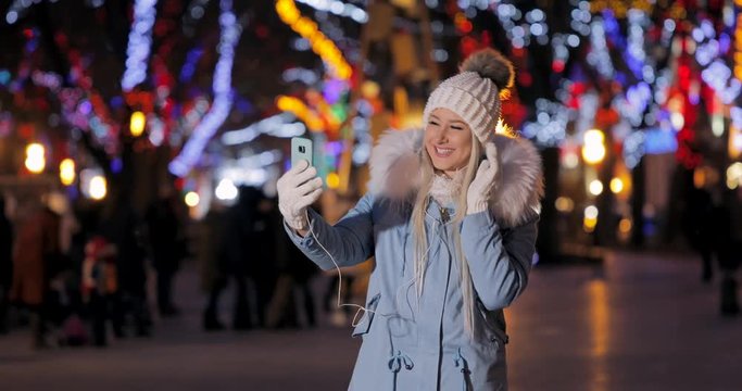 Cute and shy attractive young girl teenager takes incoming call, uses her smartphone to connect with her guy or friends, chilly winter day in the city, selfie on Christmas bright lights background.