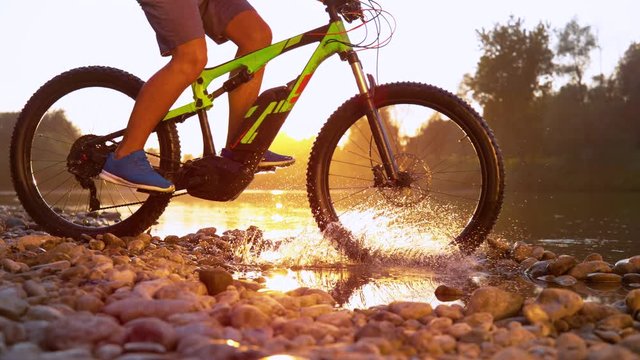 SLOW MOTION CLOSEUP Unrecognizable young man mountain biking into shallow river water at golden sunset. Mountain biker riding cross country bike over river. Freeride biking over water in sunny morning