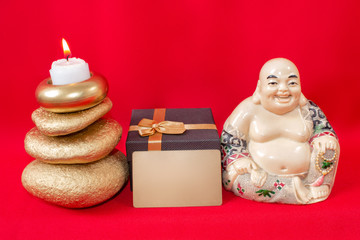  Statuette of a laughing Buddha with stones and a candle, and a gift box and a business card with free text, on a red background, feng shui.