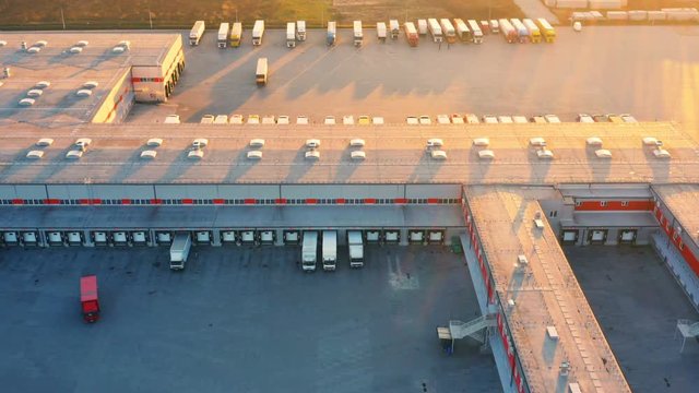 Aerial view of logistics warehouse, loading hub with many semi-trailers truck load/unload goods at sunset