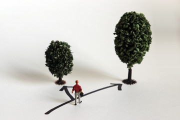 A miniature man standing in front of two trees of different sizes.