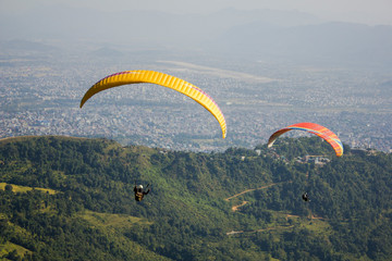 two paragliders on yellow and red parachutes on the background of green mountains and a city in the valley