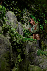 Fototapeta na wymiar Girl in a green jacket with blond hair near an artificial rock with a grotto. Fabulous photo shoot in the Studio