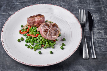 Beef medallions wrapped in bacon, serve with green peas and on a plate on wooden background. Close up