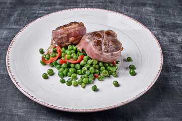 Beef medallions wrapped in bacon, serve with green peas and on a plate on wooden background. Close up