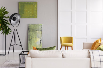 Lamp between plant and green posters in grey living room interior with chair and settee. Real photo