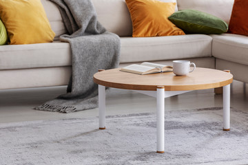 Round wooden table with book and cup on grey carpet next to sofa in flat interior. Real photo
