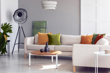 Table next to corner couch with pillows in grey living room interior with lamp and plant. Real photo