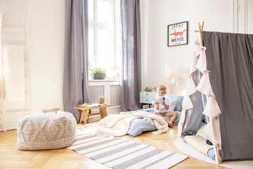 Cute boy sitting on bed in scandinavian bedroom interior with tent and big comfortable pouf, real photo