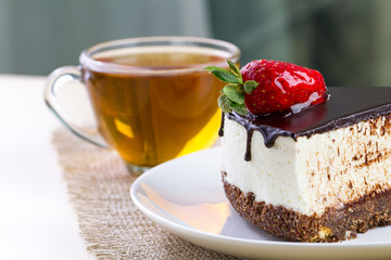 A cup of hot tea and a slice of sweet cake with whipped cream, fresh strawberries and dripping chocolate glaze in a white plate. Sweet dessert