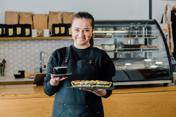 Portrait of a pretty barista greet visitors holding cup of cappuccino and server with greek cake at...