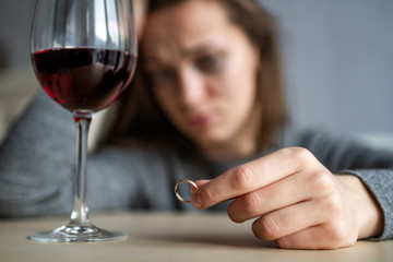 Crying, divorced woman holds a wedding ring in her hands and drinks a glass of wine because of...