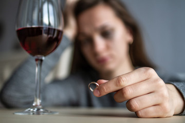 Crying, divorced woman holds a wedding ring in her hands and drinks a glass of wine because of adultery, the betrayal of her husband and a failed marriage. Divorce concept. Life problems