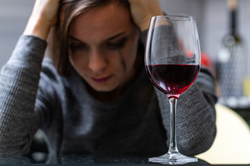 Depressed, divorced crying woman sitting alone in kitchen at home and drinking a glass of red wine because of problems at work and troubles in relationships. Social and life problems