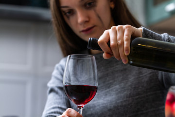 Depressed, divorced crying woman sitting alone in kitchen at home and drinking a glass of red wine because of problems at work and troubles in relationships. Social and life problems
