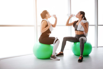 Obraz premium Two beutiful and slim young women sit on green fitballs in fitness room. They drink water from bottles. Models have rest after workout.