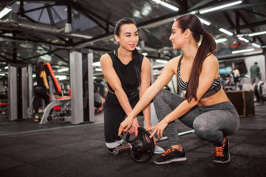 Picture of cheerful young women stand on knees togethr and smile. They look at each other. Yung asian woman help with weight for barbell.