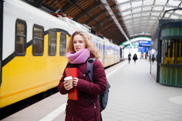A young girl stands on the platform of the station waiting for the train with a paper cup with coffee in their hands