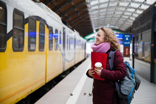 A young girl with curly hair and a backpack is standing near the train at the station and a friend to the window. Holding a paper cup of coffee and a red folder for papers
