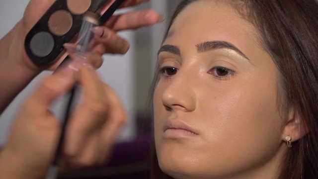 Beauty saloon. Young beautiful girl model is sitting in the chair. Makeup artist makes girl makeup. Makeup artist applies shadows on the eyelids of the model.