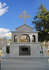 Typowy nagrobek z białego marmuru, z krzyżem i dwoma aniołami po bokach, z tyłu, na cypryjskim cmentarzu, w tle rozmyte inne nagrobki, drzewo, błekitne niebo - obrazy, fototapety, plakaty