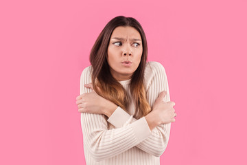 Obraz premium Dissatisfied woman feels cold during freezing chilly weather, hugs herself and clenches teeth, feels discomfort, afraids of something, isolated over pink background. Close up shot