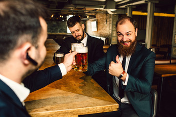 Cheerful young businessmen sit at table. They hold mugs of bear. Bearded guy hold big thumb up.