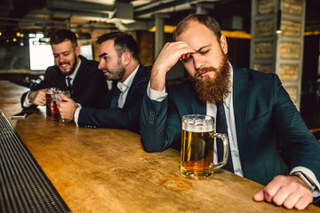 Upset and tired young man sit t bar counter. He look down and hold hand on forehead. Mug beer stand on bar counter. Other two young men sit behind.