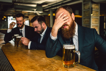 Exhausted and sad young bearded man cover face with hand. He sit at bar counter. Beer mug is there. Other two men sit behind.