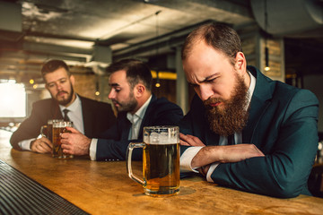 Sleepy young man sleep at bar counter. There are beer mug. Two other man sit behind and talk. They...