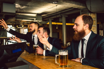 Young men in suits sit in bar and cheer. They watch TV. Guys are emotional. They wave and reach hands forward.