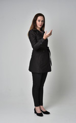 full length portrait of a brunette girl wearing long black coat, standing pose on grey studio background.