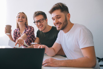 Cheerful and positive young coworkers laughing. They sit at table in white room. People look at...