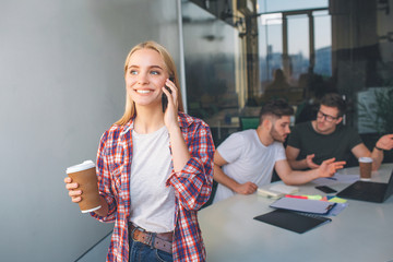 Cheerful young woman stand and talk on phone. She smiles. Model hold cup of coffee. Two young men...