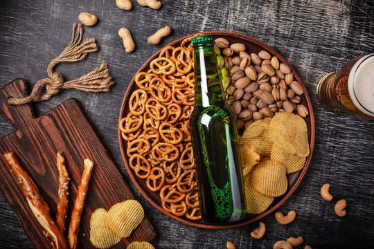 A bottle of beer on a plate with salted ookies pretzels, pistachio nuts and chips on a black scratched chalk board